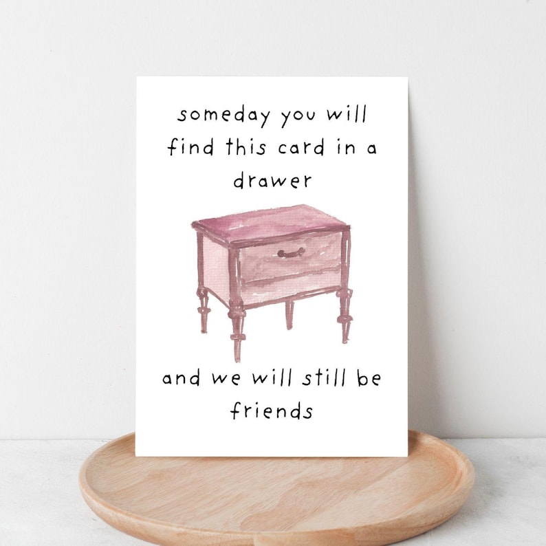 Peut inclure: Une carte de v&oelig;ux &agrave; l'aquarelle avec le texte "someday you will find this card in a drawer and we will still be friends." La carte pr&eacute;sente un dessin d'une petite table de chevet rose.