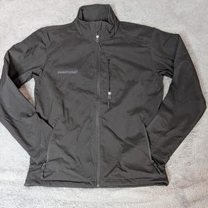May include: A black softshell jacket featuring a mock neck collar, a full-length front zipper, and long sleeves. The jacket includes two lower zippered pockets and one vertical zippered chest pocket with the text SWISSTECH printed on the right side.