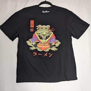May include: A black short-sleeve t-shirt featuring a graphic of a green frog wearing a purple and red patterned kimono, holding a bowl of ramen with chopsticks. The design includes red Japanese text, a red rectangular symbol, and a circular emblem.