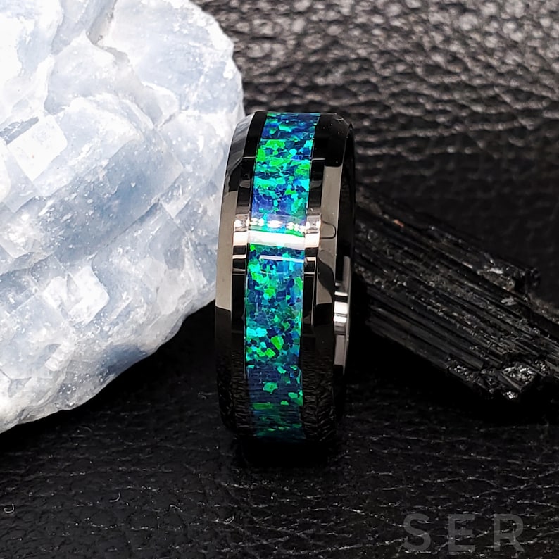 May include: A black tungsten carbide ring with a green and blue opal inlay. The ring is polished and has a beveled edge.