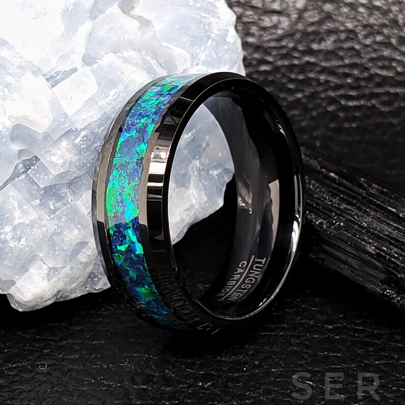 May include: Black tungsten carbide ring with a green and blue opal inlay. The ring is engraved with "TUNGSTEN CARBIDE".