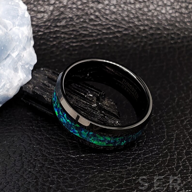 May include: Black tungsten carbide ring with a green and blue opal inlay. The ring is engraved with the words "Tungsten Carbide".