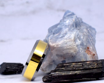 May include: A gold and silver tungsten carbide ring with a beveled edge. The ring is sitting on a blue and white crystal and a black rock.
