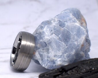 May include: A silver wedding band with two grooves around the band. The ring is sitting on a blue and white crystal rock.