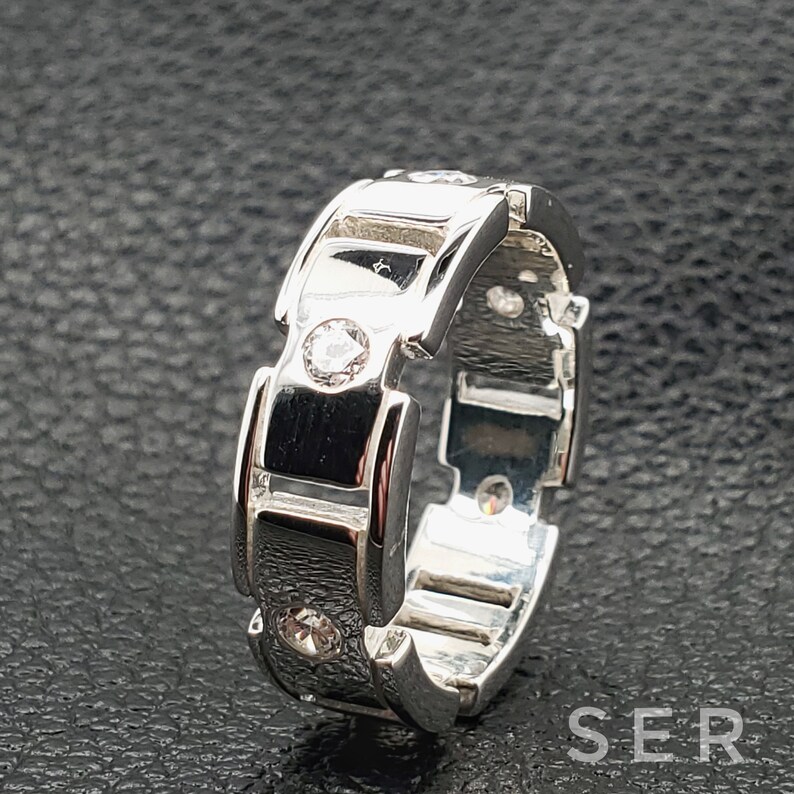 May include: A silver ring with a unique design featuring multiple small clear stones. The ring has a textured, almost chain-like design.