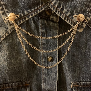 Pode incluir: Clipes de colarinho prateados em forma de planeta com três correntes penduradas, presos a uma camisa jeans. O design do planeta tem um anel ao redor da esfera. A camisa é de ganga de lavagem média.