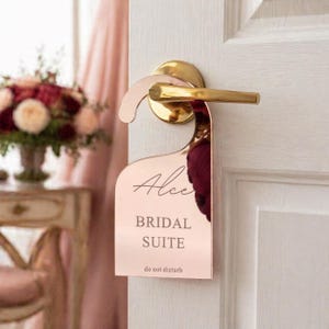 Könnte beinhalten: Ein roségoldener Türhänger mit dem Namen "Alice" in Schreibschrift, den Worten "BRIDAL SUITE" und "do not disturb". Der Hänger hängt an einer weißen Tür mit einem goldenen Griff. Im Hintergrund ist ein Blumenarrangement zu sehen.