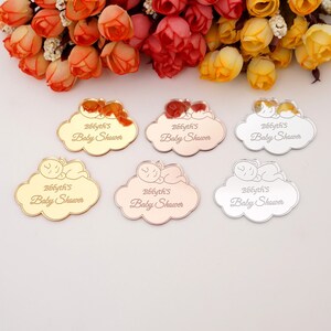 May include: Six cloud-shaped baby shower favors in gold, rose gold, and silver. Each favor features a sleeping baby design and the text "Bblyth's Baby Shower" in a script font. The favors are displayed against a backdrop of colorful flowers.