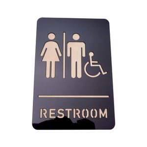 May include: Black restroom sign with cut-out icons for male, female, and wheelchair accessible. The word "RESTROOM" is cut out at the bottom of the sign. The sign is rectangular with rounded corners.