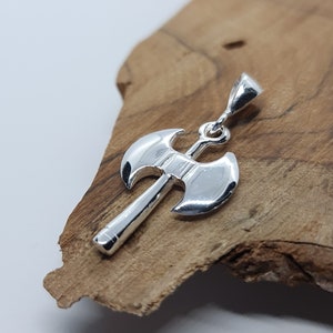 May include: A silver axe pendant with a detailed double-bladed head and a long handle. The pendant hangs from a small loop. The axe is displayed on a piece of light brown wood.