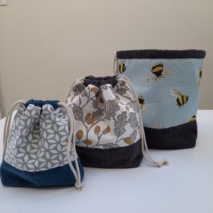 May include: Three drawstring bags with different patterns. The largest bag is blue with a white background and a bee pattern. The middle bag is white with a grey background and a leaf pattern. The smallest bag is grey with a white background and a geometric pattern.