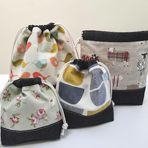 May include: Four drawstring bags in different sizes. The bags display diverse fabric patterns, including birds, sheep, and floral designs. Each bag has a black denim base and a drawstring closure with a white cord.
