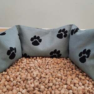 Puede incluir: Tres cojines de almohada azul claro con dise&ntilde;os de huellas de patas negras. Los cojines est&aacute;n dispuestos en semic&iacute;rculo sobre un lecho de c&aacute;scaras de almendras marrones.