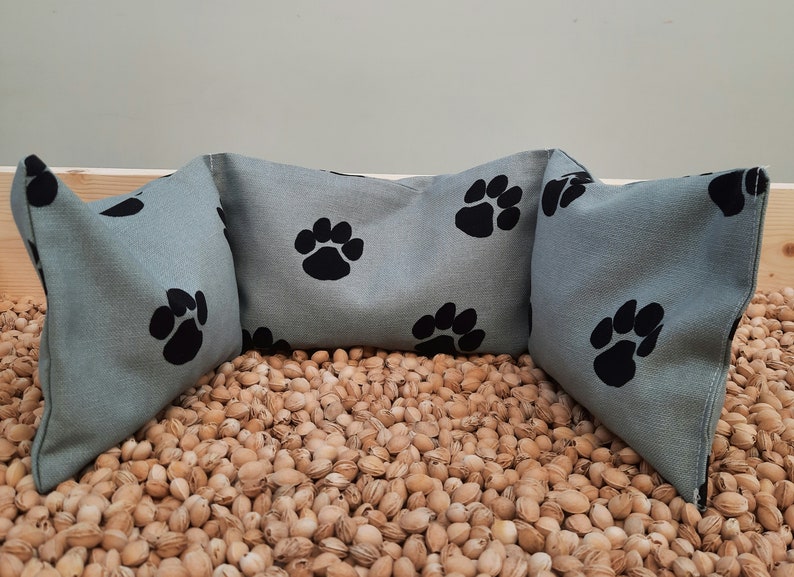 Puede incluir: Tres cojines de almohada azul claro con dise&ntilde;os de huellas de patas negras. Los cojines est&aacute;n dispuestos en semic&iacute;rculo sobre un lecho de c&aacute;scaras de almendras marrones.
