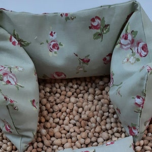 Puede incluir: Una almohada verde claro con un patr&oacute;n floral de rosas rosas y flores blancas. La almohada est&aacute; llena de peque&ntilde;as semillas marrones y tiene forma de U.