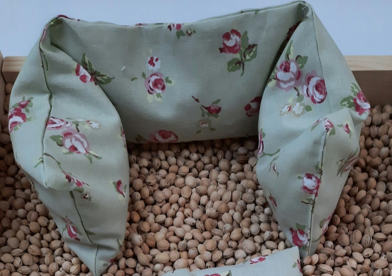 Puede incluir: Una almohada verde claro con un patr&oacute;n floral de rosas rosas y flores blancas. La almohada est&aacute; llena de peque&ntilde;as semillas marrones y tiene forma de U.