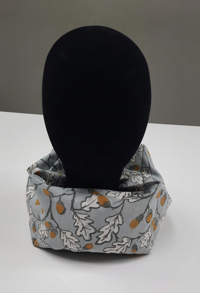 Puede incluir: Un calentador de cuello de tela gris y blanco con un patr&oacute;n de hojas de roble y bellotas. El calentador de cuello est&aacute; en una cabeza de maniqu&iacute; negra.