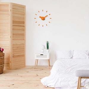 Può includere: Un orologio da parete arancione moderno con un design minimalista. L'orologio ha lancette e indicatori delle ore arancioni su una parete bianca. L'orologio ha un diametro di circa 30 cm. La stanza ha un letto, un comodino e un paravento in legno.