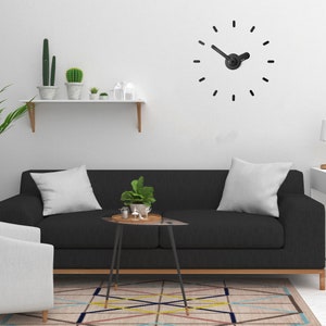 May include: A black sofa with two white pillows sits in front of a white wall with a black clock. A small black side table with a plant and decorative items sits in front of the sofa. A patterned rug is on the floor.