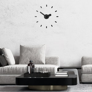 Può includere: Un orologio nero con un design minimalista è appeso a una parete bianca sopra un divano grigio con cuscini. Un tavolino da caffè nero con un piano in vetro si trova davanti al divano.