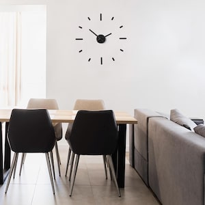 Op de afbeelding: Een zwarte minimalistische wandklok met lange, dunne wijzers en een zwarte middelste cirkel. De klok is gemonteerd op een witte muur boven een eettafel met vier stoelen. De tafel is gemaakt van hout en heeft een lichtbruine afwerking. De stoelen zijn bekleed met een donkergrijze stof.