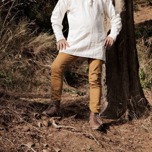 Op de afbeelding: Een persoon draagt een off-white tuniek met lange mouwen, een knoopsluiting en een bruine broek. De persoon staat naast een boom in een natuurlijke omgeving en draagt bruine laarzen.