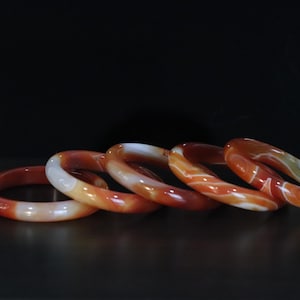 May include: A set of five circular bangles crafted from polished carnelian stone. Each bangle features a smooth, rounded surface with natural color variations ranging from deep burnt orange to translucent white, creating unique swirling patterns.