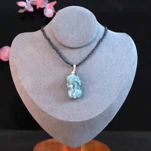 Puede incluir: Collar con colgante azul-verdoso con una figura tallada en un cord&oacute;n negro. El colgante se exhibe sobre un busto gris sobre un fondo negro. Se ven flores rosas en el fondo.