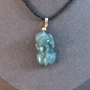 Puede incluir: Colgante de jade de color verde azulado con forma de figura sentada, suspendido de un cord&oacute;n negro. El colgante tiene detalles dorados. El fondo es gris apagado. Joyer&iacute;a.