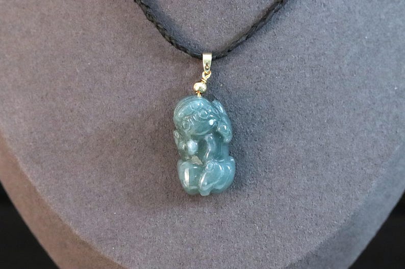 Puede incluir: Colgante de jade de color verde azulado con forma de figura sentada, suspendido de un cord&oacute;n negro. El colgante tiene detalles dorados. El fondo es gris apagado. Joyer&iacute;a.