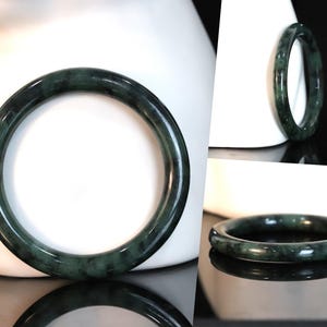 Puede incluir: Un brazalete de jade verde oscuro. El brazalete es liso y pulido, con un sutil patrón de mármol.