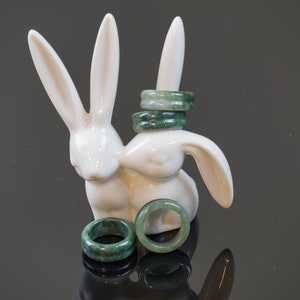 May include: Four jade green rings stacked on a white ceramic rabbit figurine. Two more jade green rings are lying on a black surface in front of the rabbit.
