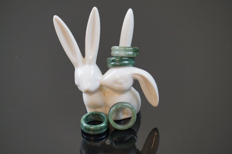 May include: Four jade green rings stacked on a white ceramic rabbit figurine. Two more jade green rings are lying on a black surface in front of the rabbit.