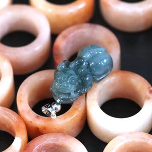 Puede incluir: Una colecci&oacute;n de anillos de jade en tonos melocot&oacute;n y naranja, con un colgante detallado de color verde azulado que presenta una figura tallada. Los anillos son lisos y pulidos, mostrando variaciones de color naturales. El colgante tiene una fianza plateada.