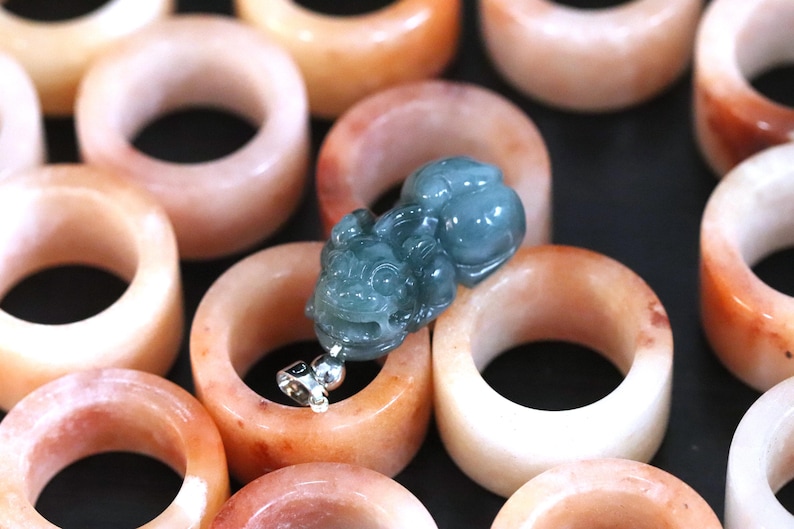 Puede incluir: Una colecci&oacute;n de anillos de jade en tonos melocot&oacute;n y naranja, con un colgante detallado de color verde azulado que presenta una figura tallada. Los anillos son lisos y pulidos, mostrando variaciones de color naturales. El colgante tiene una fianza plateada.