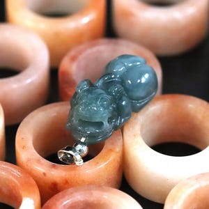 Puede incluir: Una colecci&oacute;n de anillos de jade color melocot&oacute;n con un colgante de jade verde azulado detallado. El colgante es una figura tallada con una base de metal plateado. Los anillos son lisos y pulidos, con variaciones de color.