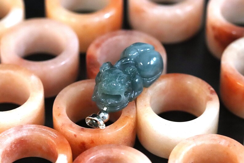 Puede incluir: Una colecci&oacute;n de anillos de jade color melocot&oacute;n con un colgante de jade verde azulado detallado. El colgante es una figura tallada con una base de metal plateado. Los anillos son lisos y pulidos, con variaciones de color.
