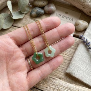 Op de afbeelding: Twee gouden kettingen met stervormige hangers. Eén hanger is heldergroen, de andere lichtgroen. De kettingen worden op een hand getoond, met een neutrale achtergrond.