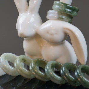 May include: A set of jade rings in various shades of green, some with a lighter, almost white hue. The rings are arranged in a semi-circle on a surface, with two white ceramic rabbit figurines in the background.