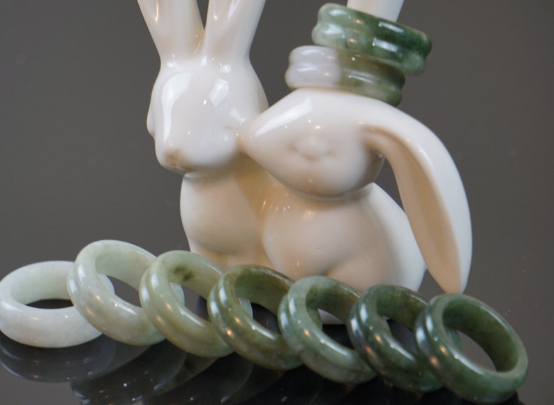 May include: A set of jade rings in various shades of green, some with a lighter, almost white hue. The rings are arranged in a semi-circle on a surface, with two white ceramic rabbit figurines in the background.