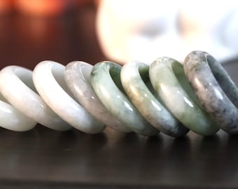 May include: A row of smooth, polished stone rings arranged in a gradient from opaque white to translucent pale green and mottled dark grey. Each ring features a rounded, thick band design with a glossy finish, showcasing natural color variations.