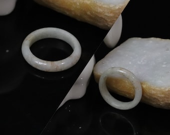 May include: Two smooth, pale green jade rings with subtle white and tan inclusions. The rings have a rounded, polished band profile and a translucent appearance, displayed against a high-contrast black and white split background.