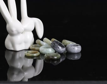 May include: A collection of jade rings in various shades of green and gray, arranged on a black surface. The rings are displayed on a white ceramic rabbit figurine.