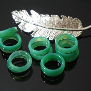 Peut inclure: Plusieurs bagues vertes translucides, peut-être en jade ou en agate, sont disposées sur une surface noire réfléchissante. Un ornement argenté en forme de plume est en arrière-plan. Les bagues sont empilées à différentes hauteurs.