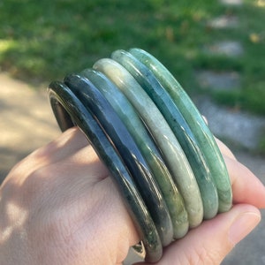 May include: A collection of jade bangles in varying shades of green, from deep forest to pale seafoam. The smooth, polished surfaces reflect light, showcasing the natural variations in color and texture. The bangles are stacked together, displaying their circular form.