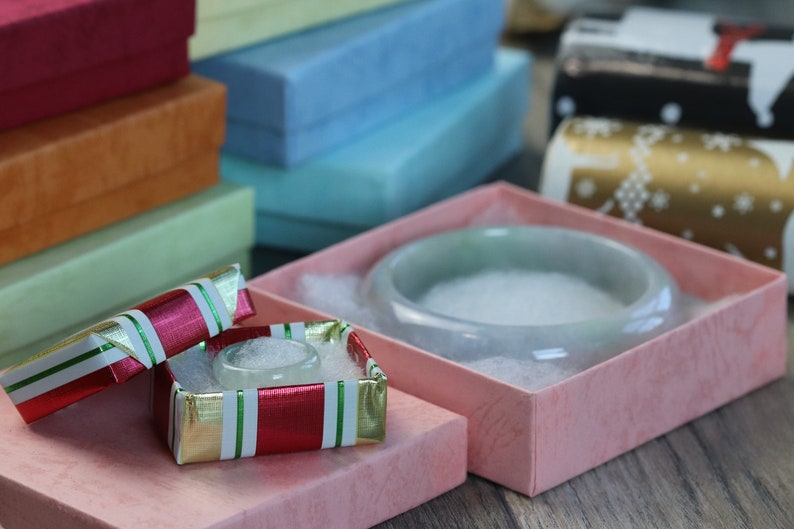 May include: Two small gift boxes with lids open. The box on the left is red and white with green trim. The box on the right is pink with a white interior and a white ceramic bowl inside.