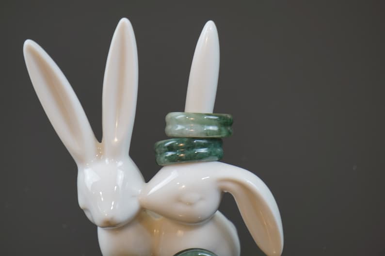 May include: Three jade rings stacked on a white ceramic rabbit figurine. The rings are a light green colour with darker green veins.