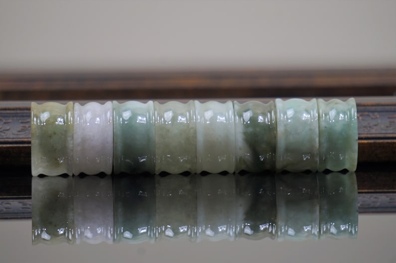 May include: A set of seven jade green and white jade rings with a ridged design. The rings are arranged in a row on a dark surface.