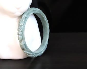 May include: A pale green jade bangle bracelet with carved floral and dragon motifs. The bracelet is resting on a white surface with a black background. The jade is a translucent, light green color.