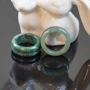 May include: Two jade green rings, one with a smooth surface and the other with a textured surface, are displayed on a white ceramic rabbit figurine.